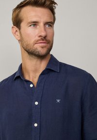 Navy blue linen shirt features a button-down collar, white buttons, and a small embroidered design on the chest. Smooth texture, relaxed fit.