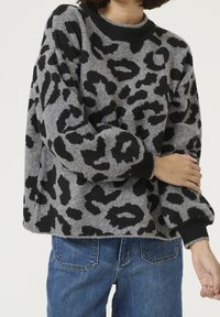 Pull en tissu gris avec un imprimé léopard noir. Il présente un col rond et des manches longues, avec des poignets et un ourlet côtelés noirs. Jeans visible.