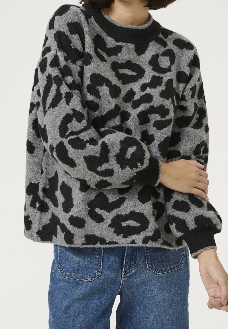 Pull en tissu gris avec un imprimé léopard noir. Il présente un col rond et des manches longues, avec des poignets et un ourlet côtelés noirs. Jeans visible.