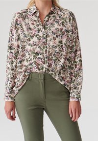 Blouse fluide à fleurs en crème, ornée de fleurs roses et vertes, à fermeture boutonnée et à manches longues. Associée à un pantalon slim fit vert olive.