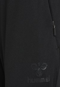 Zbliżenie na czarną tkaninę z czarnym suwakiem i wytłoczonym logo pszczoły Hummel na odzieży sportowej.