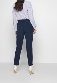 Kvinna klädd i marinfärgade skräddarsydda byxor, vitrandig blus och vita högklackade ankelstövlar, som står med ryggen mot kameran på en enfärgad bakgrund.