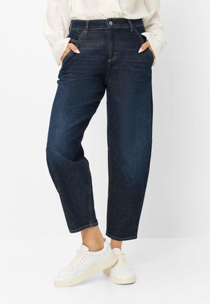 Kvinde iført mørkeblå jeans med høj talje og lige ben, hænderne i lommerne, kombineret med hvide sneakers og en cremefarvet bluse.