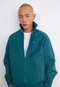 Casaco azul-turquesa com fecho éclair e gola alta, apresentando punhos elásticos e o logótipo da Adidas. Textura suave com bolsos laterais para funcionalidade adicional.