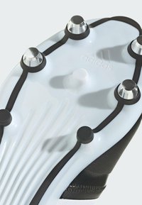 Podešev kopaček Adidas na fotbal, která má bílý základ s černými akcenty, několik stříbrných kolíků a texturované drážky pro lepší trakci.