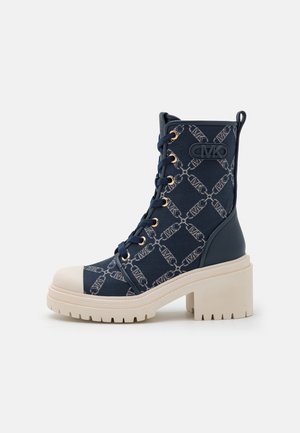 Botte à lacets bleu marine avec talon épais beige et bout renforcé, tissu à motifs avec chaîne et design de logo, et œillets dorés.