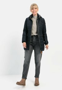 Sort, quiltet pufferjakke med hætte og frontlynlås, parret med en lys grå kabelstrikket sweater og mørke jeans. Brune støvler.