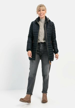 Zwarte, gewatteerde pufferjas met een capuchon en een ritssluiting aan de voorkant, gecombineerd met een lichtgrijze kabelgebreide trui en donkere jeans. Bruine laarzen.