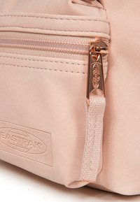 Ljusrosa Eastpak-väska med en texturerad yta, synlig dragkedja och en metallflik. Präglat logotyp på framsidan visar varumärket.