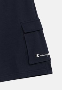 Σκούρο μπλε ύφασμα με μία τσέπη καπάκι και λευκό κεντημένο λογότυπο "Champion" στην κάτω δεξιά γωνία ενός ρούχου.