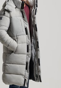 Parka acolchada gris con cierre de cremallera y broches, acentos negros y capucha, con un diseño texturizado y acolchado y bolsillos laterales.