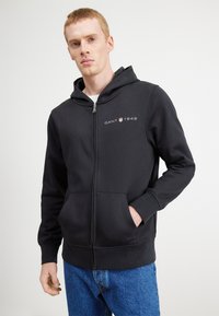 Felpa nera con zip realizzata in tessuto morbido, dotata di tasca frontale e cappuccio. Presenta un piccolo logo "GANT 1949" stampato sul petto.
