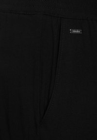 Černé látkové kalhoty s boční kapsou, jemným švem a malým kovovým logem blízko pásku. Hladká textura.