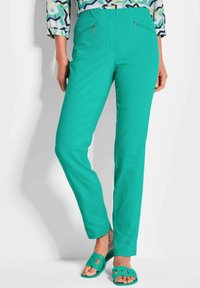 Türkise Hose mit geradem Schnitt, ausgestattet mit vorderen Reißverschlusstaschen und einem glatten, dehnbaren Stoff. Kombiniert mit grünen Sandalen.