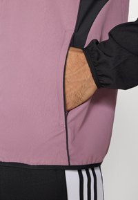 Ροζ ανοιχτόχρωμο μπουφάν με μαύρες λεπτομέρειες, που διαθέτει πλαϊνή τσέπη και ελαστικές μανσέτες, κατασκευασμένο από ένα λείο, ελαφρύ ύφασμα.