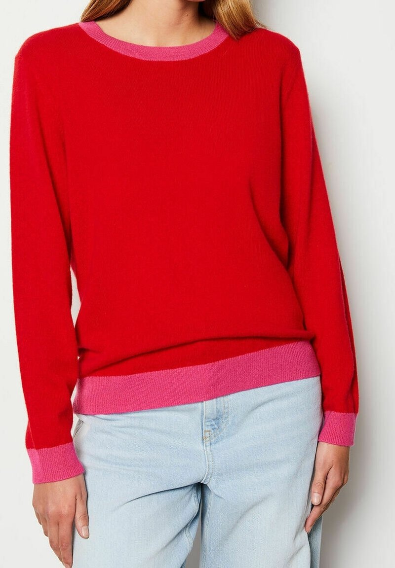 Femme portant un pull rouge vif avec des bordures roses au col, aux poignets et à l'ourlet, associé à un jean bleu clair.
