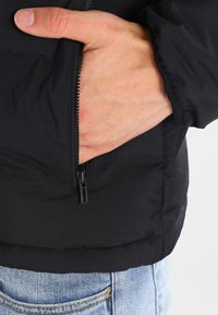 Veste matelassée noire avec fermeture éclair à l'avant, dotée d'une poche latérale. Texture lisse et poignets élastiques. Portée avec un jean bleu clair.