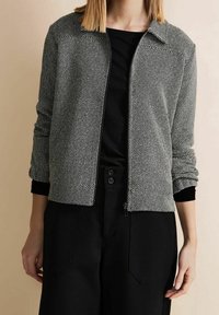 Szara i biała kurtka z zamkiem błyskawicznym z wzorem, z kołnierzykiem i czarnymi mankietami, w połączeniu z czarną bluzką i luźnymi czarnymi spodniami.