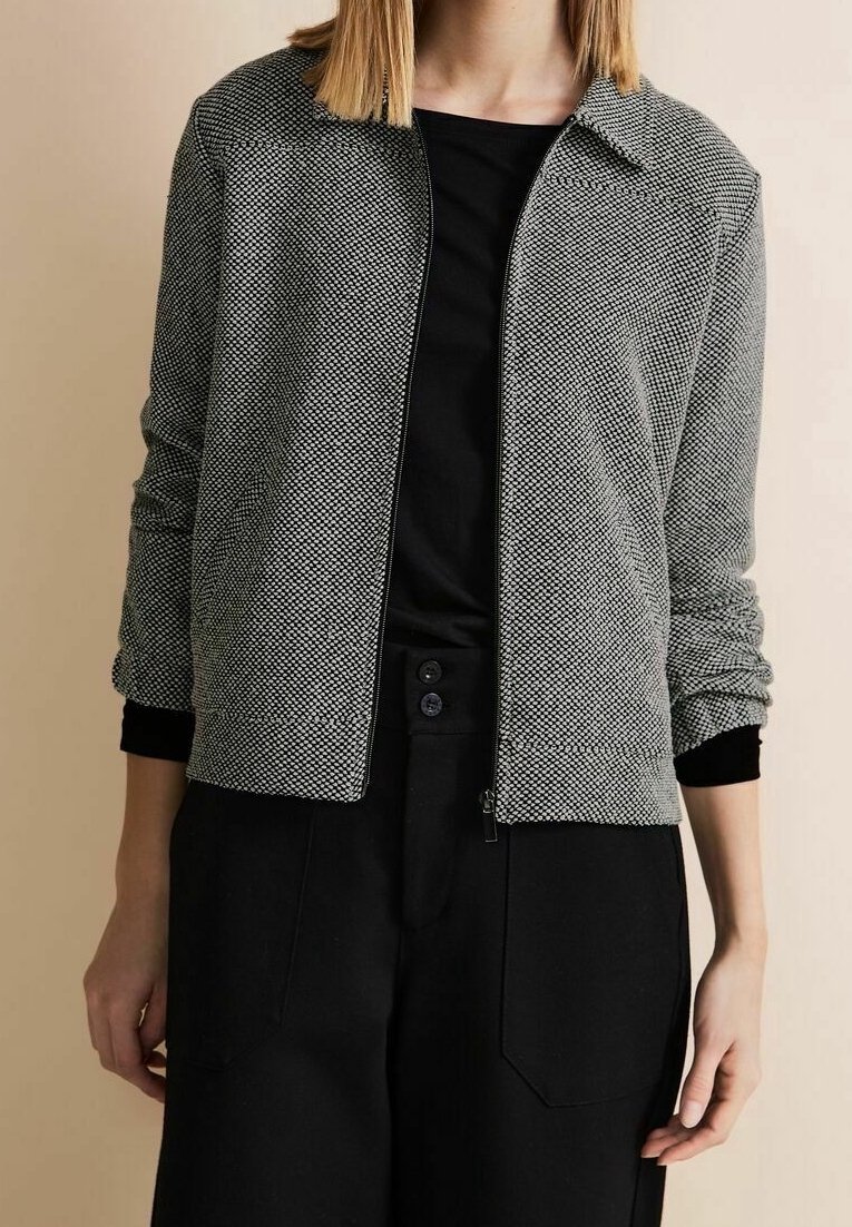 Szara i biała kurtka z zamkiem błyskawicznym z wzorem, z kołnierzykiem i czarnymi mankietami, w połączeniu z czarną bluzką i luźnymi czarnymi spodniami.