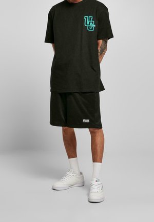 T-shirt en coton noir avec un logo turquoise sur la poitrine, associé à un short noir assorti. Des baskets blanches et des chaussettes mi-mollet sont portées.