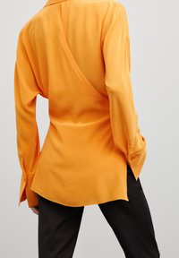 Oranges Langarmhemd mit asymmetrischem Rückendesign, aus glattem Stoff, mit geknöpften Manschetten und abgerundetem Saum.