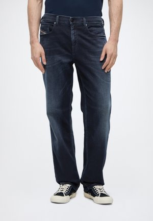 Hombre con vaqueros rectos azul oscuro y zapatillas negras con cordones, de pie frente a un fondo claro liso.