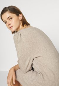 Abito lavorato a maglia beige con vestibilità comoda, scollo rotondo e maniche a sbuffo. Texture morbida con lievi variazioni di colore.