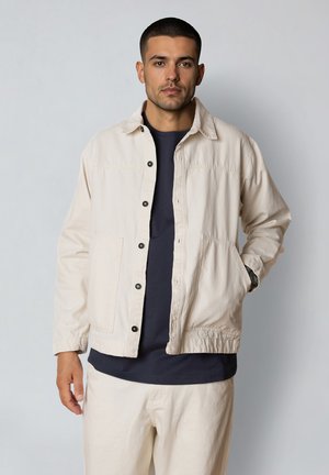 Man met kort donker haar, gekleed in een lichtbeige jas met knopen over een marineblauw shirt en bijpassende beige broek, staand tegen een effen achtergrond.