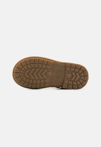 Suela de goma con un patrón de tracción texturizado, que cuenta con ranuras y crestas para un mejor agarre. Color: marrón claro. Forma plana y redondeada adecuada para diversos terrenos.