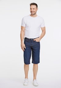 Shorts de mezclilla azul oscuro con un corte relajado, cuentan con acentos cosidos y bolsillos delanteros, combinados con una camiseta blanca ajustada y zapatillas blancas.