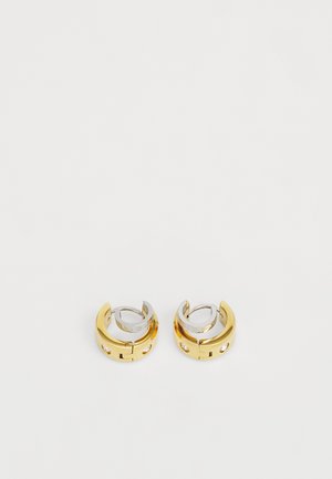 Guld- och silverfärgade metallhänge i form av ringar med en elegant design, som har en slät yta och små cirkulära utskärningar.