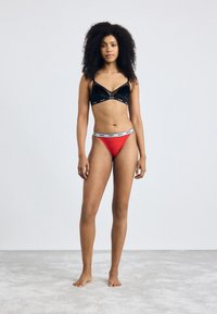 Black velvet bralette with a triangular design and logo accent; red thong with a contrasting logo waistband; model standing on a light gray surface.