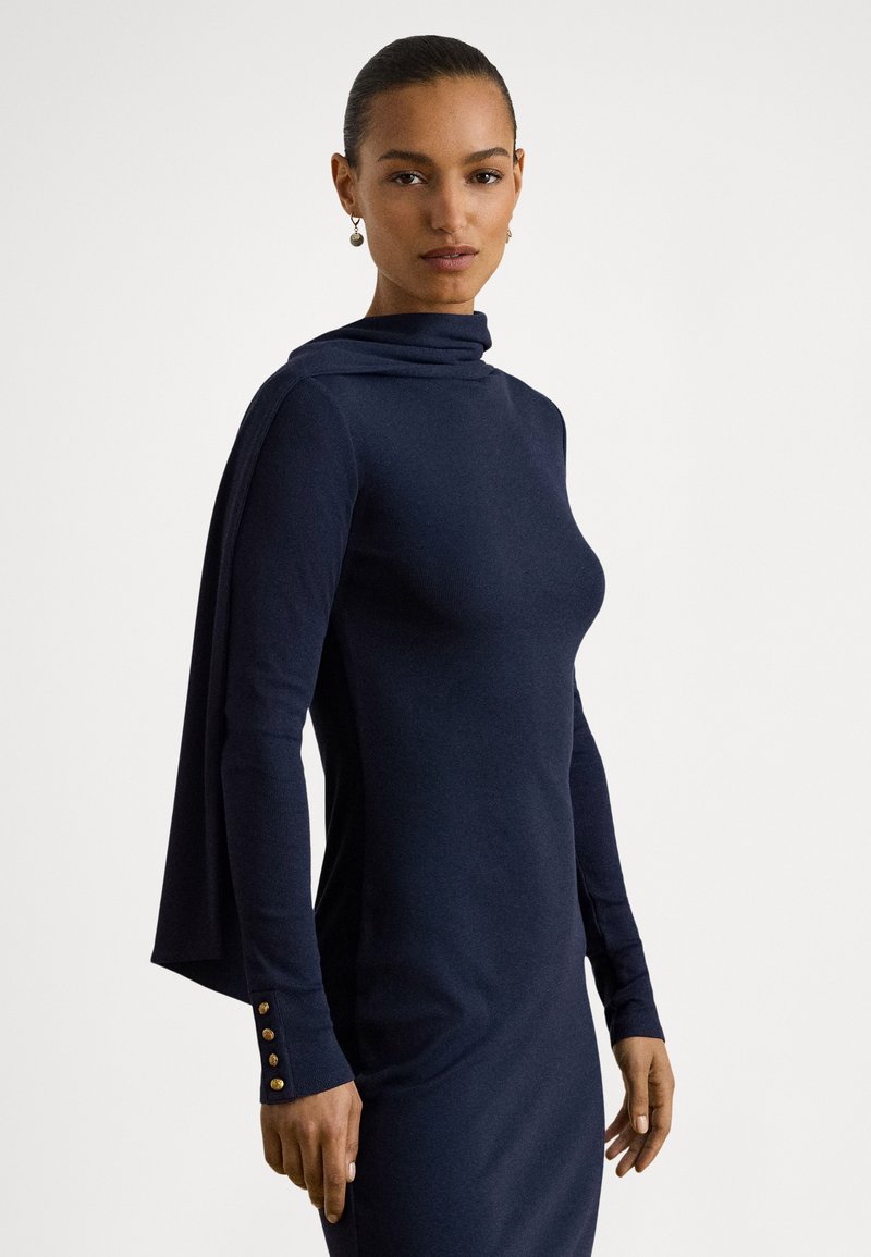 Tamsiai mėlyna suknelė su aukštu kaklu, ilgomis rankovėmis ir apsiausto detalėmis. Rankogaliuose puošta aukso spalvos sagutėmis. Pagaminta iš minkštos, tekstūruotos medžiagos.