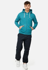 Haut à zip en teal avec une capuche doublée, des cordons de serrage et des poches avant, associé à un pantalon sombre et des baskets blanches.