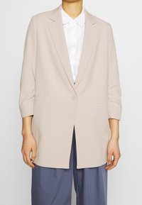 Blazer beige avec une fermeture à un seul bouton, des revers crantés, des manches trois-quarts et des poches latérales, associé à une chemise blanche et un pantalon bleu.