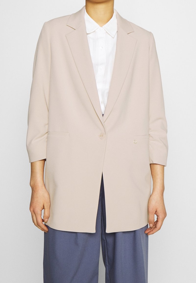 Blazer beige avec une fermeture à un seul bouton, des revers crantés, des manches trois-quarts et des poches latérales, associé à une chemise blanche et un pantalon bleu.