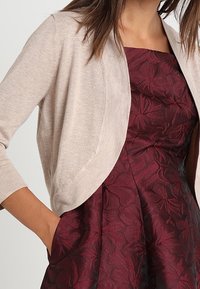 Beige cardigan with 3/4 sleeves, lightweight knit material, open front design, paired with a deep burgundy dress featuring floral patterns.
