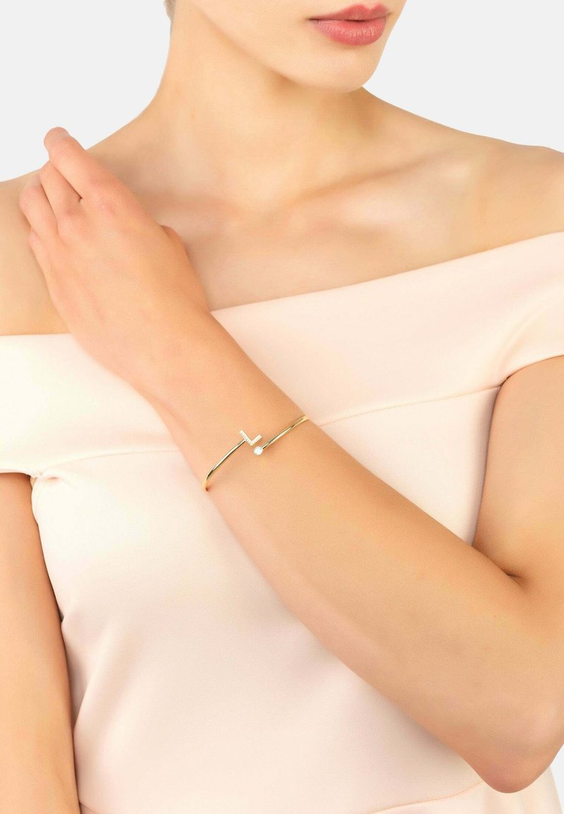 Gold bangle with a minimalist design, featuring a geometric accent and a small decorative stone. Worn on a bare arm against a soft background.
