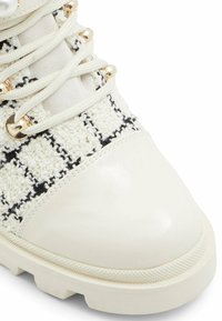 Białe sneakersy z teksturowaną, plecioną cholewką z czarnym wzorem w kratę, błyszczącą, białą osłoną palców oraz złotymi akcentami na sznurówkach.
