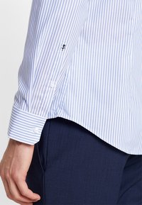 Světle modrá a bílá pruhovaná košile s manžetou na knoflík, na rukávu s květinovou výšivkou a se zaobleným lemem.