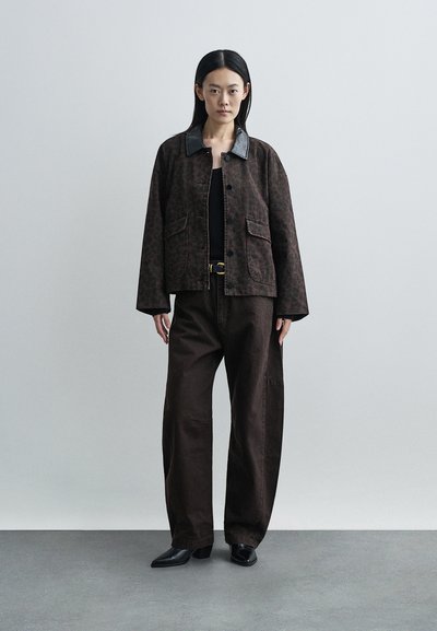 Casaco castanho estampado com gola preta, combinado com calças largas castanho escuro e sapatos pretos. Modelo está em pé contra um fundo simples.