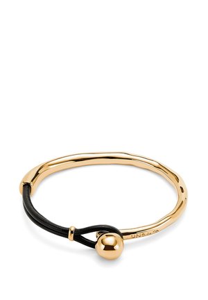 Pulsera de metal en tono dorado con un diseño suave y curvado, que presenta un cordón de cuero negro y un detalle de cierre esférico.