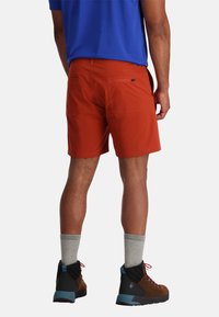 Pantaloni corti rossi da uomo con due tasche posteriori, realizzati in tessuto leggero. Abbinati a stivali da escursionismo marroni e calze grigie.