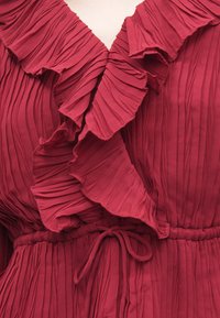 Blusa plissada cor burgundy com decote em V com folhos e cintura franzida com laço. Tecido leve com acabamento texturado.