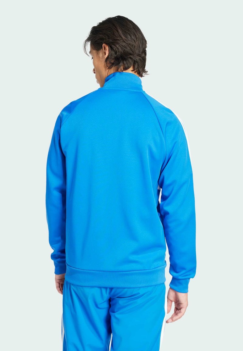 Niebieska sportowa kurtka z wysokim kołnierzem, prążkowanymi mankietami i pasem. Posiada gładką teksturę oraz kontrastowe białe paski po bokach.