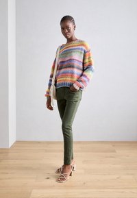Multikleurige gebreide trui met horizontale strepen, olijfgroene slim-fit broek en witte strappy hakken. Vlak geweven textuur, casual pasvorm.