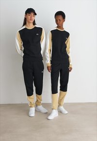 Tute da allenamento neri e beige; maniche lunghe, pannelli a contrasto sulle spalle e pantaloni in stile jogger. Entrambi indossano sneakers bianche.