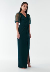 Garas zaļš kleita ar V-veida kakla izgriezumu un savītā priekšpuses dizainu. Caurspīdīgas puff piedurknes ar pērlīšu akcentiem un sānu šķēlumu. Gluda auduma tekstūra.