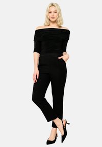 Črn top brez naramnic z nabranim materialom, v kombinaciji z utesnjenimi črnimi hlačami in čevlji s koničastim nosom, kar ustvarja eleganten silhueto.
