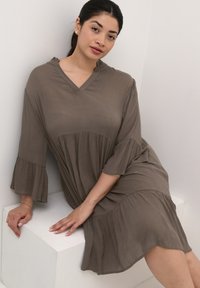Robe brune à col en v avec des manches évasées et une silhouette fluide, faite d'un tissu texturé. Modèle assis, les mains reposant sur une surface blanche.